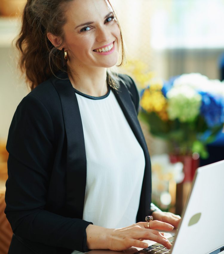 smiling elegant woman in white blouse and black jacket at modern home in sunny day using website on a laptop while sitting on couch. typing on keyboard